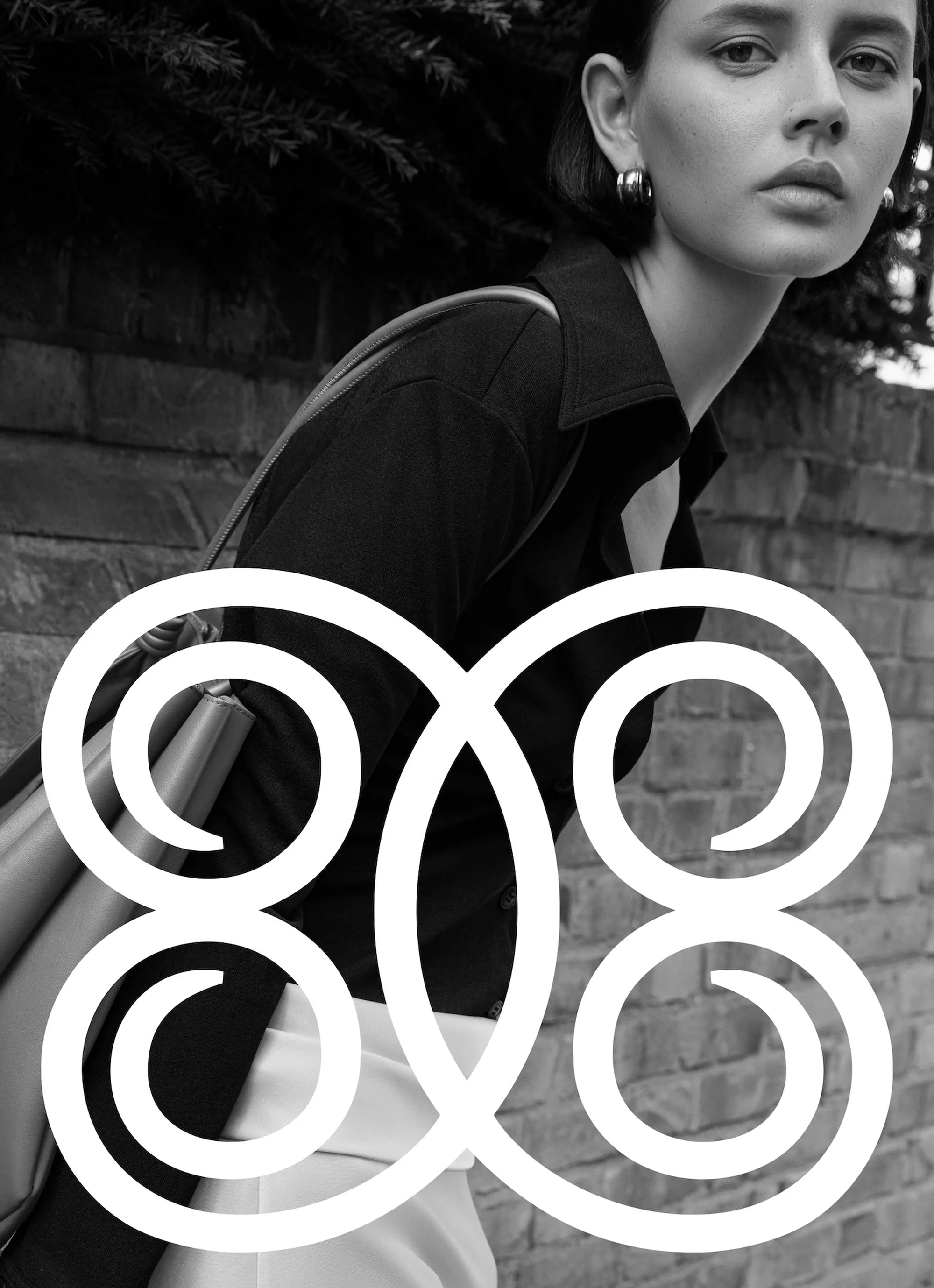 Black and white portrait of a woman in a black shirt, silver hoop earrings, with a handbag, posing in front of a brick wall.