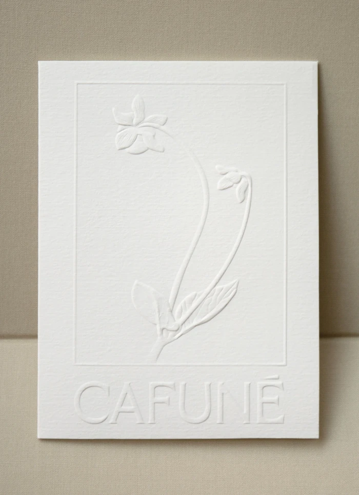White embossed paper card featuring a botanical floral illustration and Cafune brand text