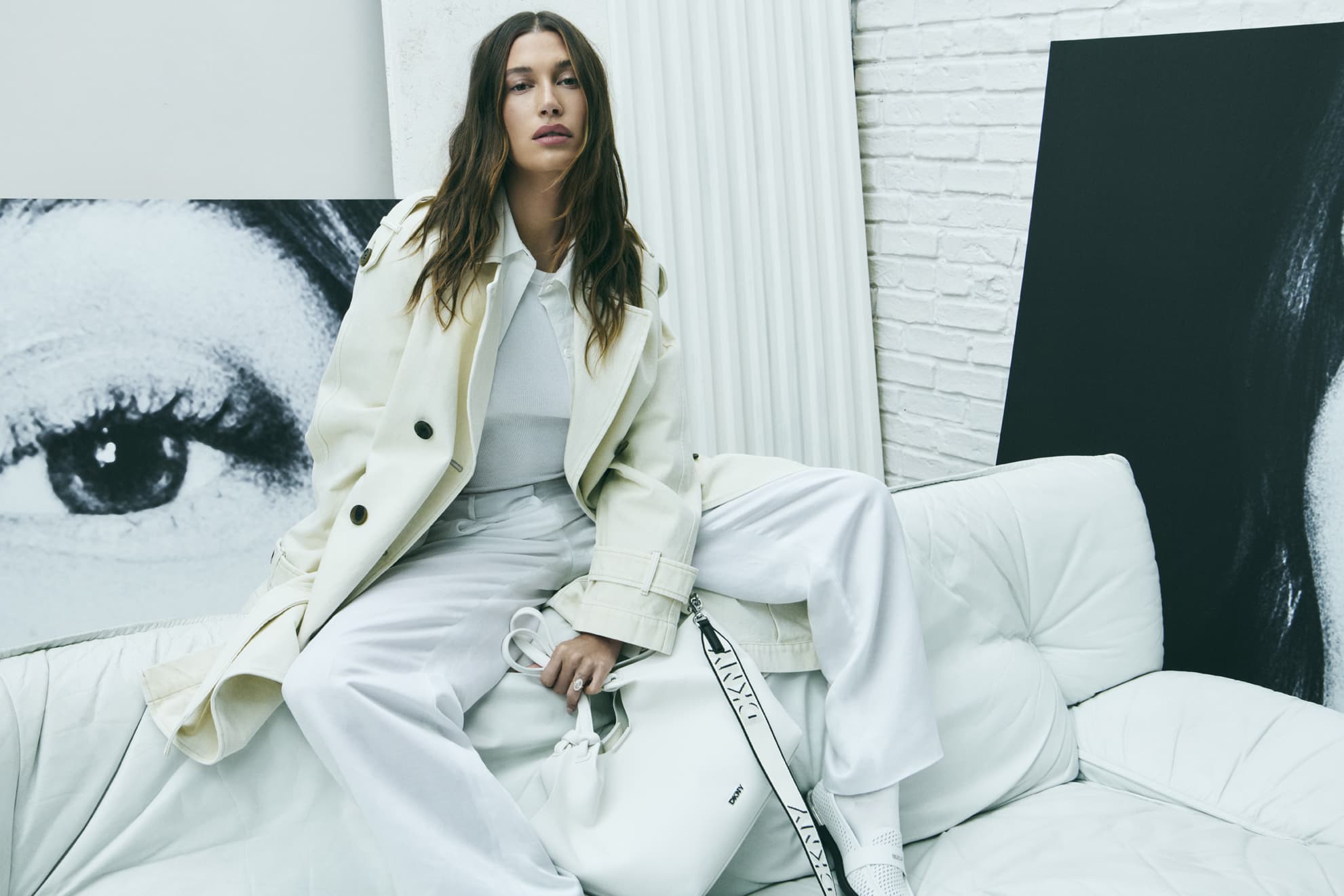 A female fashion model in an off-white coat and pants sits on a white sofa next to a large poster in a neutral indoor setting.
