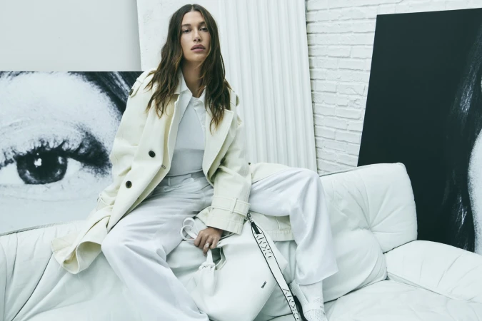 A female fashion model in an off-white coat and pants sits on a white sofa next to a large poster in a neutral indoor setting.