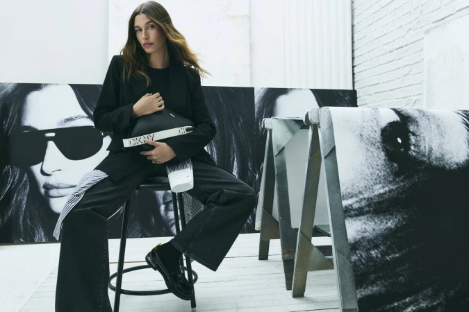 Adult female in a black blazer and dark denim poses with a black DKNY purse in a photo studio with backdrop portrait prints.