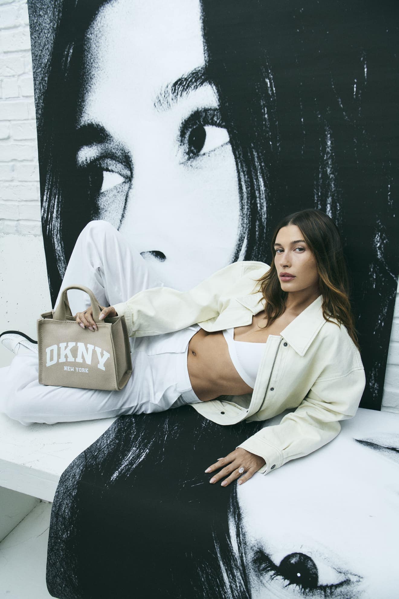 Hailey Bieber poses with a DKNY bag in front of a black-and-white portrait backdrop, wearing a white crop top and a light yellow jacket.