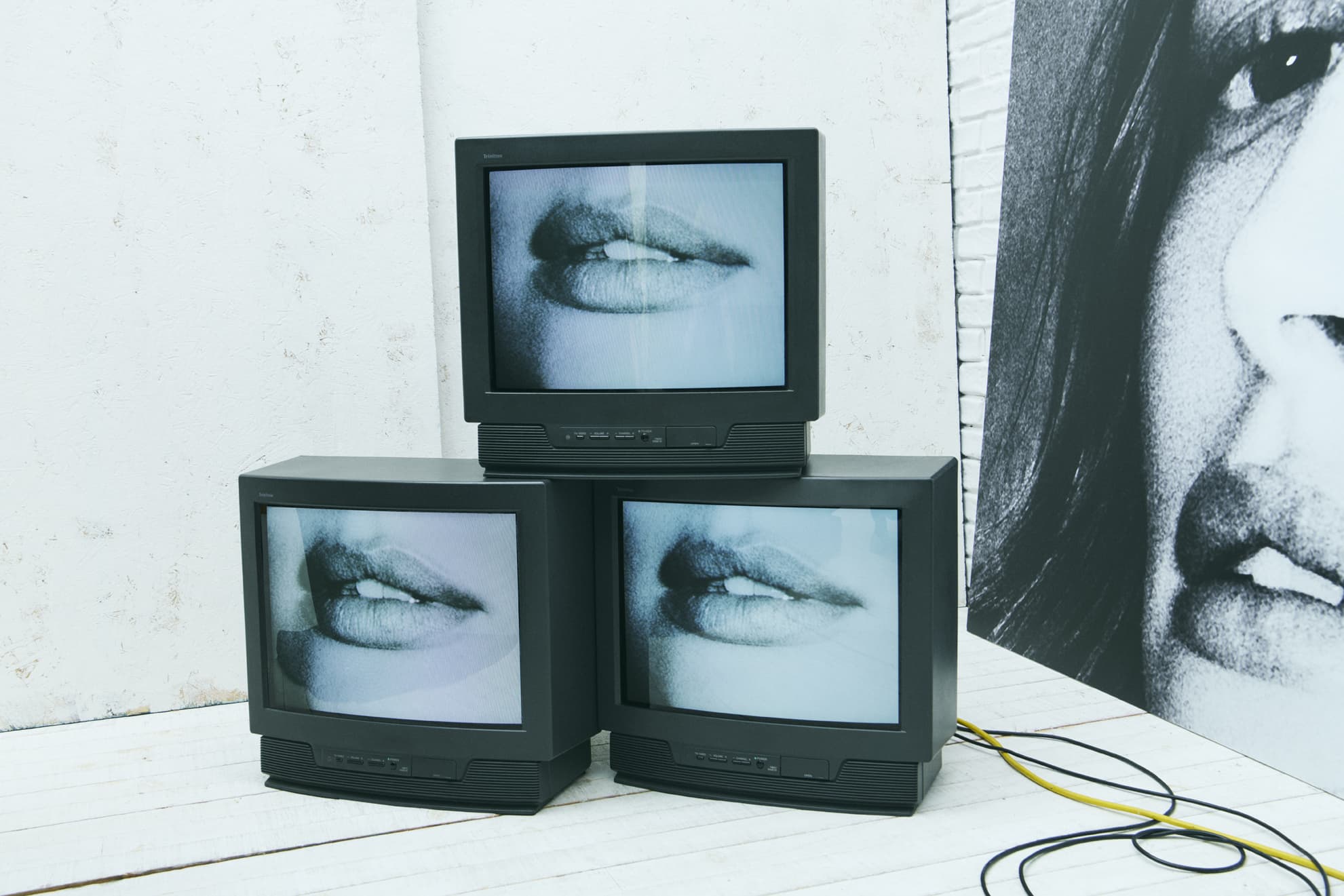 Three vintage CRT televisions stacked, displaying a close-up of open lips against a white brick wall with hanging wires and a poster.