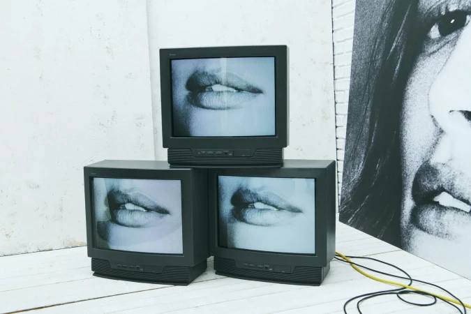 Three vintage CRT televisions stacked, displaying a close-up of open lips against a white brick wall with hanging wires and a poster.