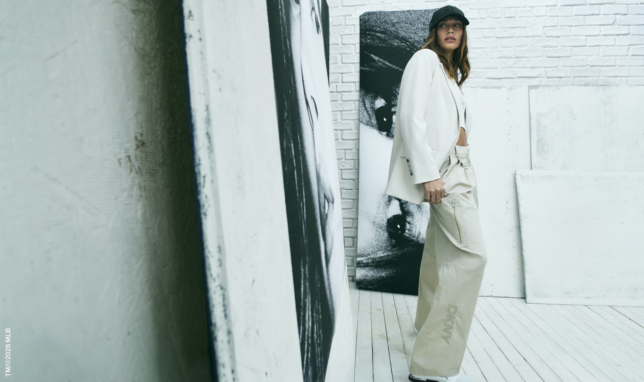 A fashionable woman poses in a white blazer and beige pants, standing against backdrop of black and white art and a white brick wall.