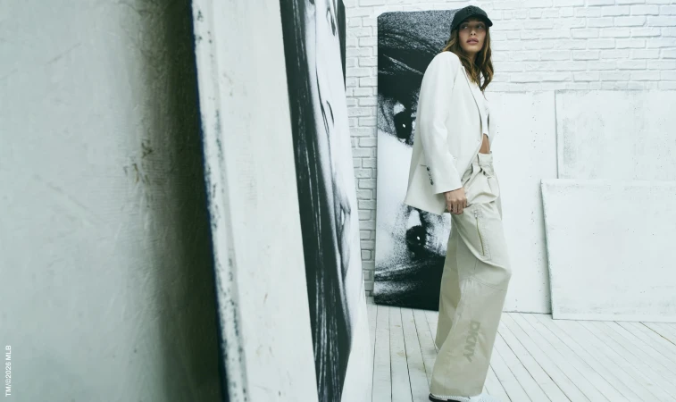A fashionable woman poses in a white blazer and beige pants, standing against backdrop of black and white art and a white brick wall.