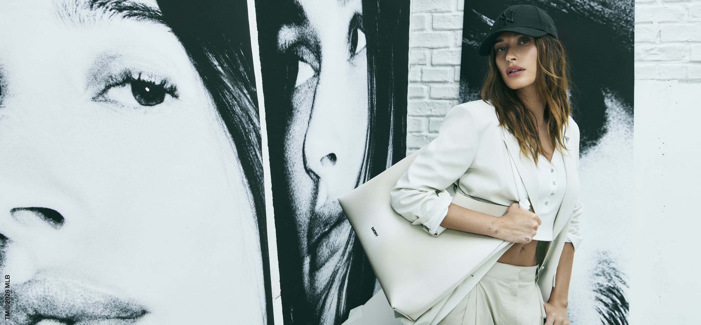 Woman in a white blazer poses with a tote bag in front of a black and white mural and a white brick wall indoors.