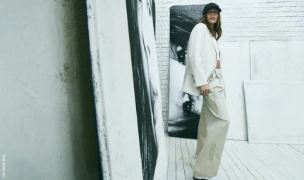Hailey Bieber wearing an off-white blazer and cream trousers with a black cap in a white studio setting.