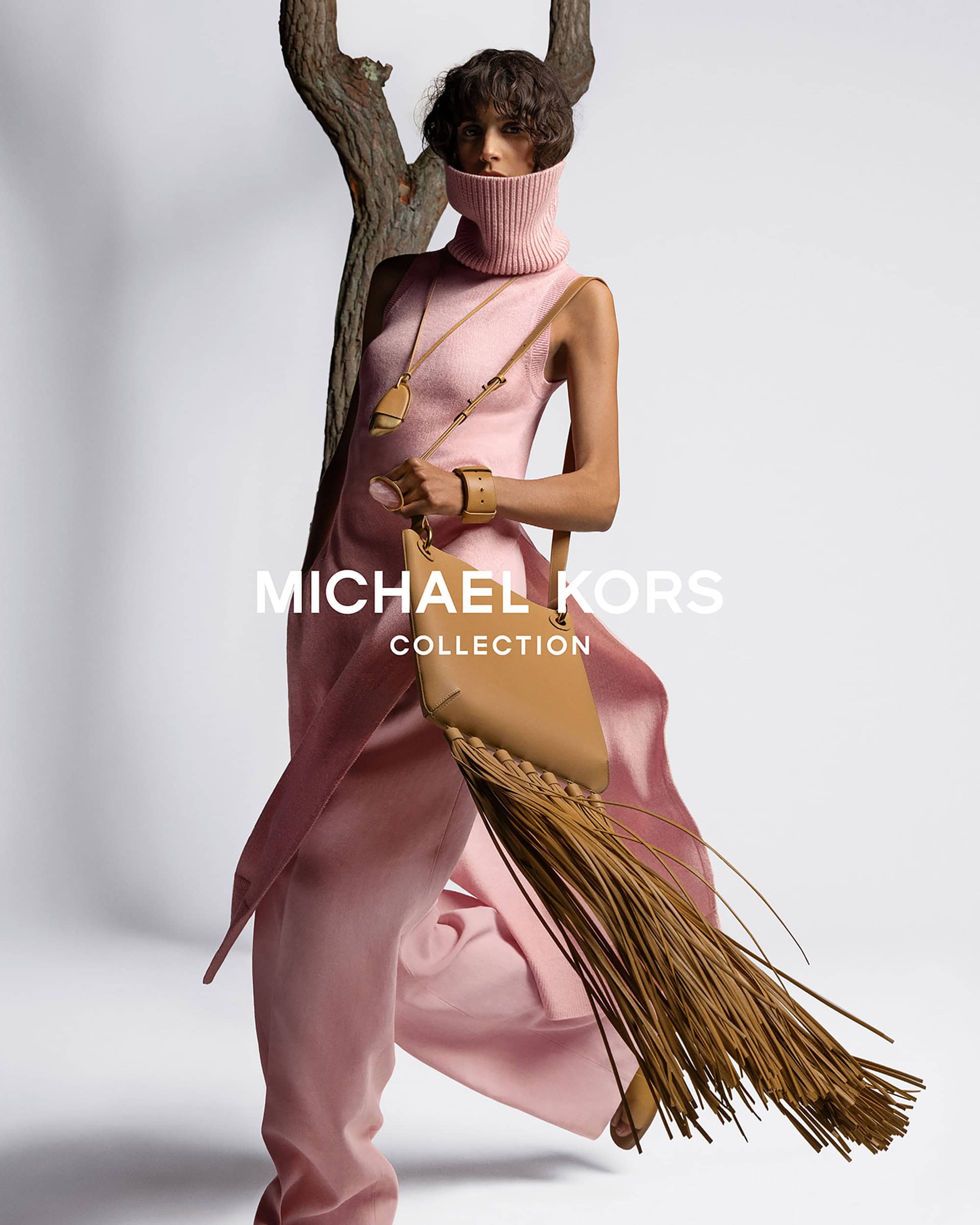 Fashion shot of a model in a pink turtleneck outfit, accessorized with a leather handbag with fringe, against a neutral backdrop.