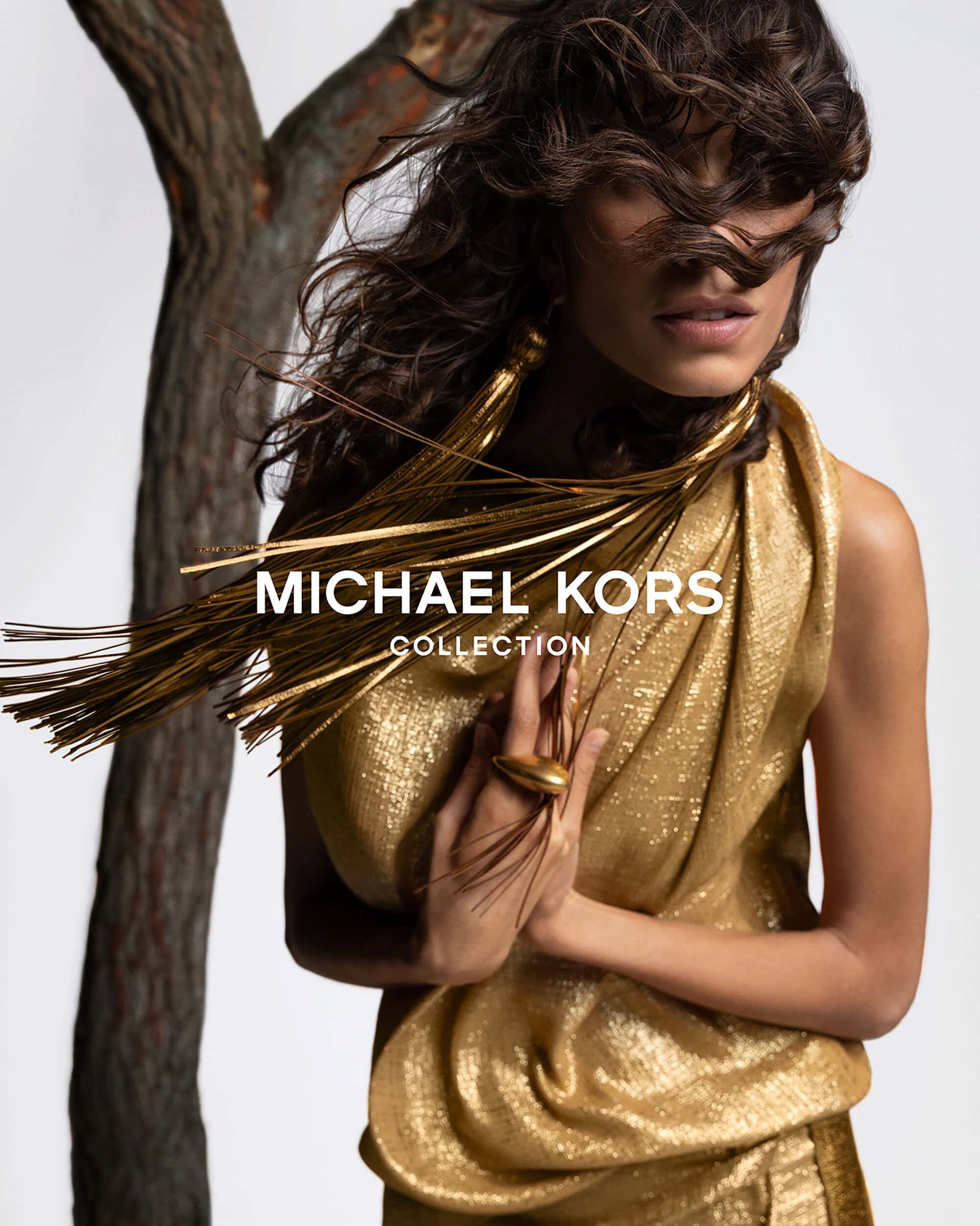 A woman models a gold, shimmering dress and tassel jewelry with a blurred tree branch and logo in the background in a fashion shot.