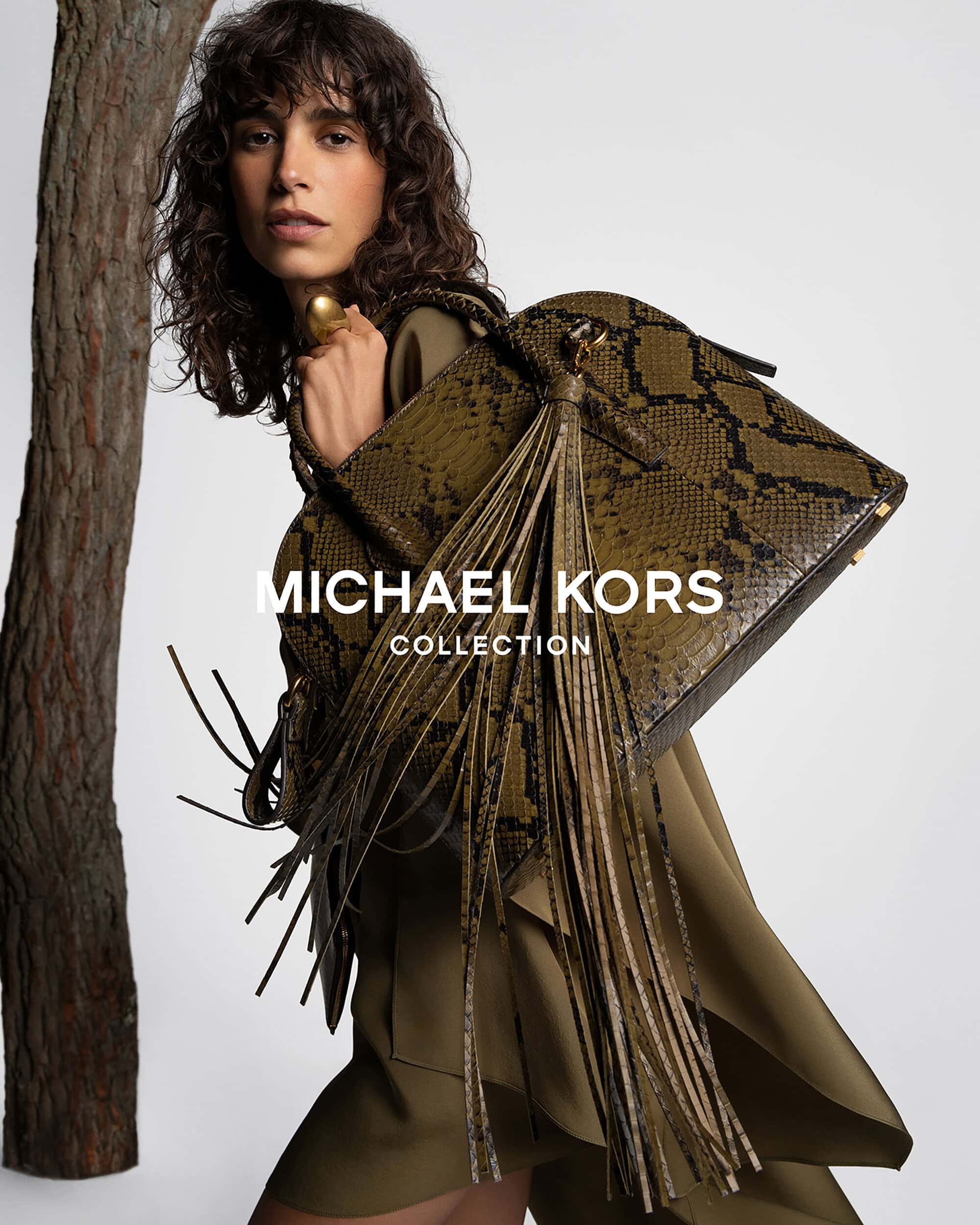Fashion shot featuring a woman with curly hair wearing a green outfit, holding a snakeskin print handbag next to a tree trunk.