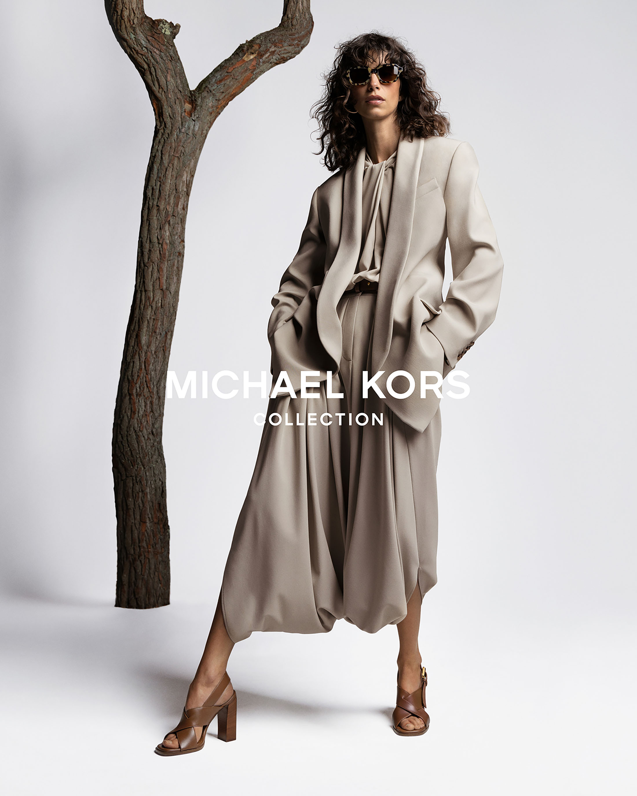 Model Mica Argañaraz wearing a tailored beige suit and sunglasses in a studio ad campaign featuring a stylized tree prop.