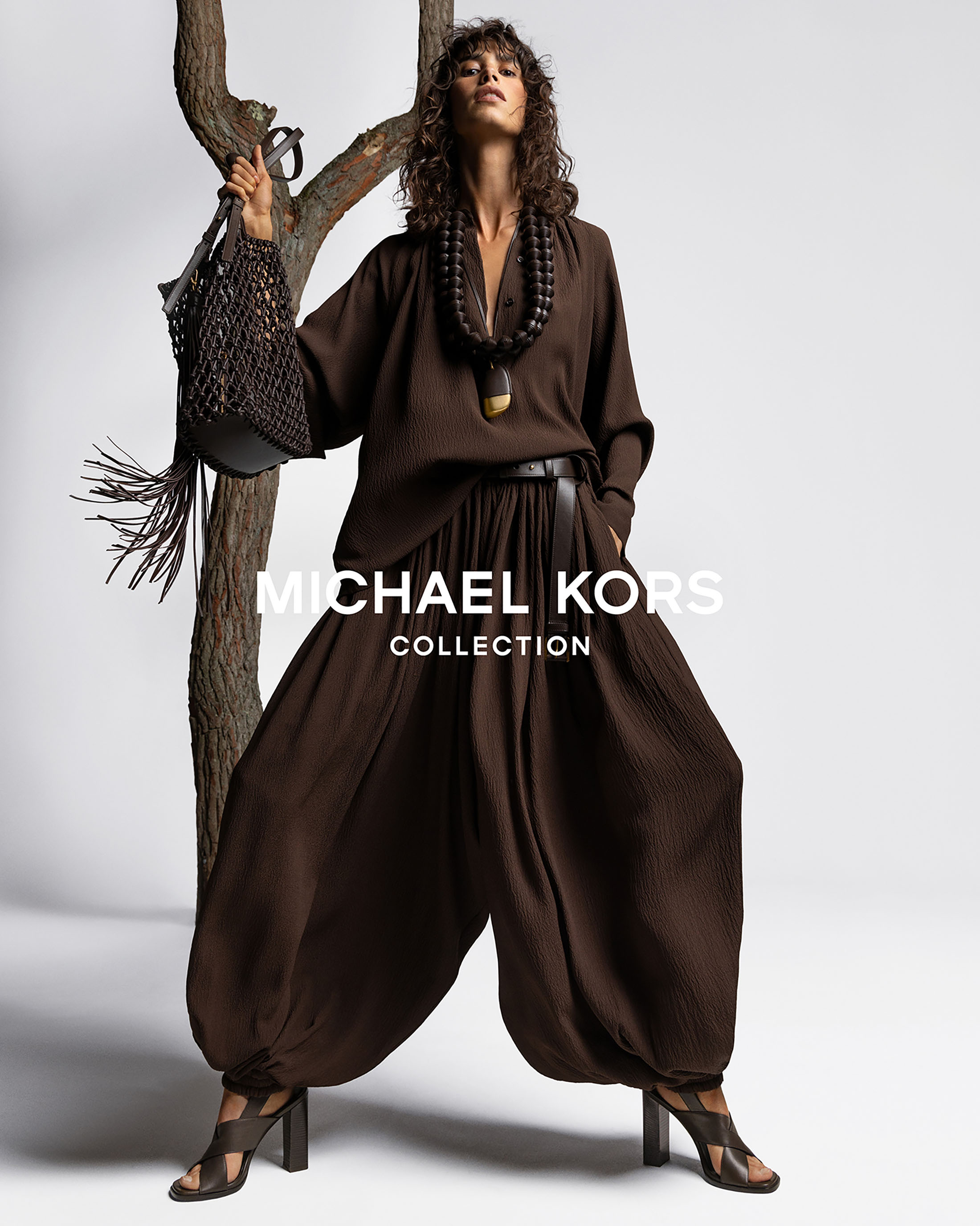 Fashion model in brown Michael Kors outfit with slouchy pants and necklace poses next to a tree branch on a white background.