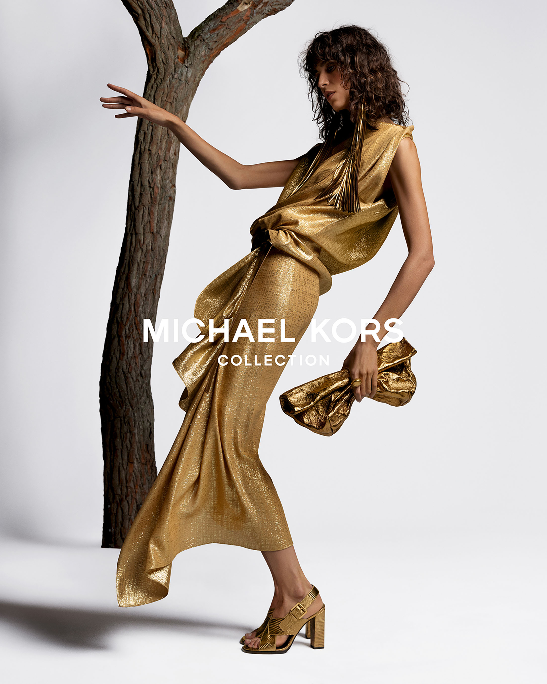 Fashion model wears a glittering gold dress and strappy heels, posing near a tree branch for a Michael Kors Collection fashion ad.