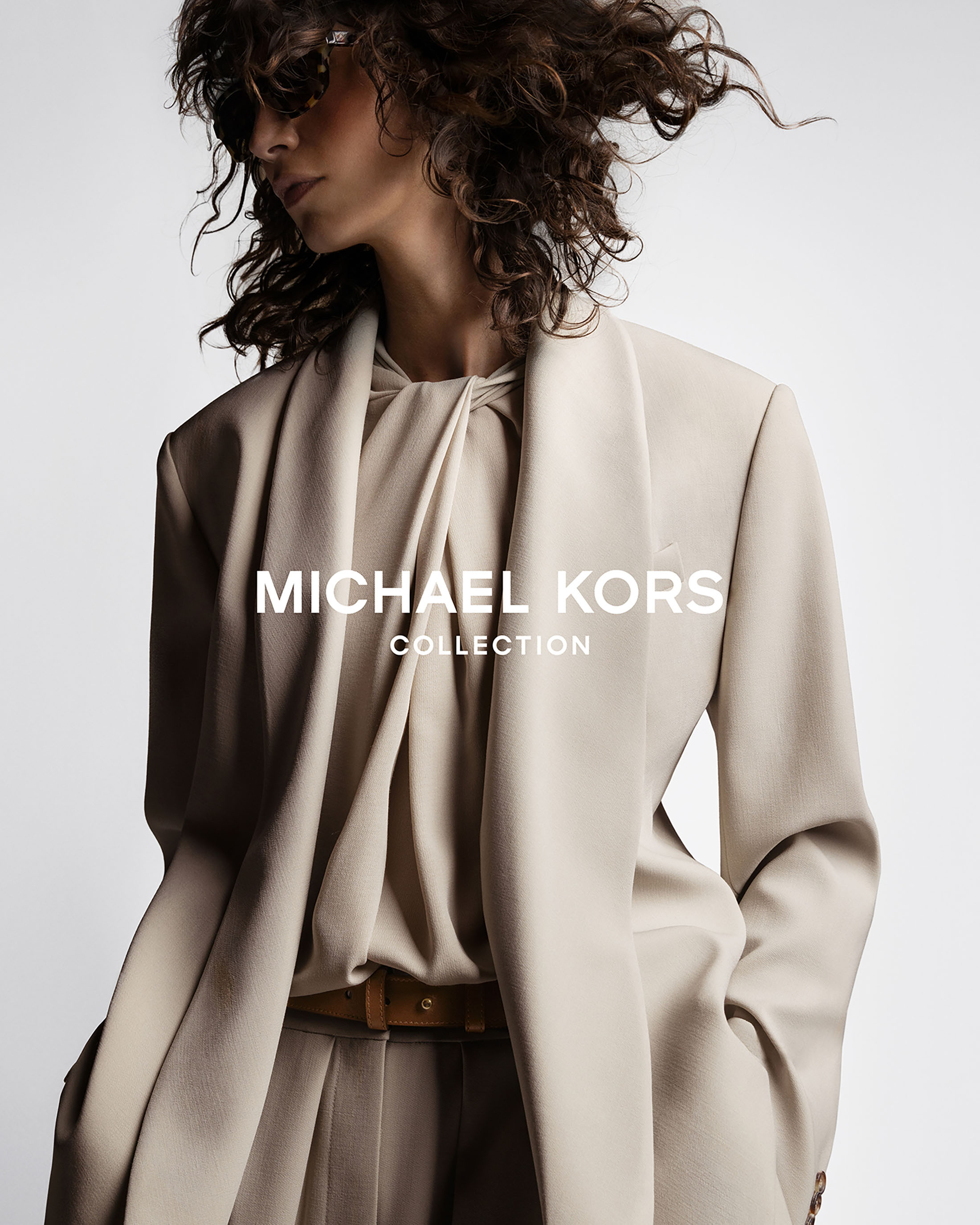 Fashion advertisement featuring a woman wearing a beige blazer and pleated trousers by Michael Kors Collection against a white backdrop.