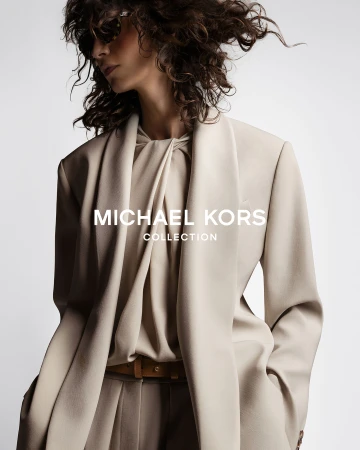 Fashion advertisement featuring a woman wearing a beige blazer and pleated trousers by Michael Kors Collection against a white backdrop.