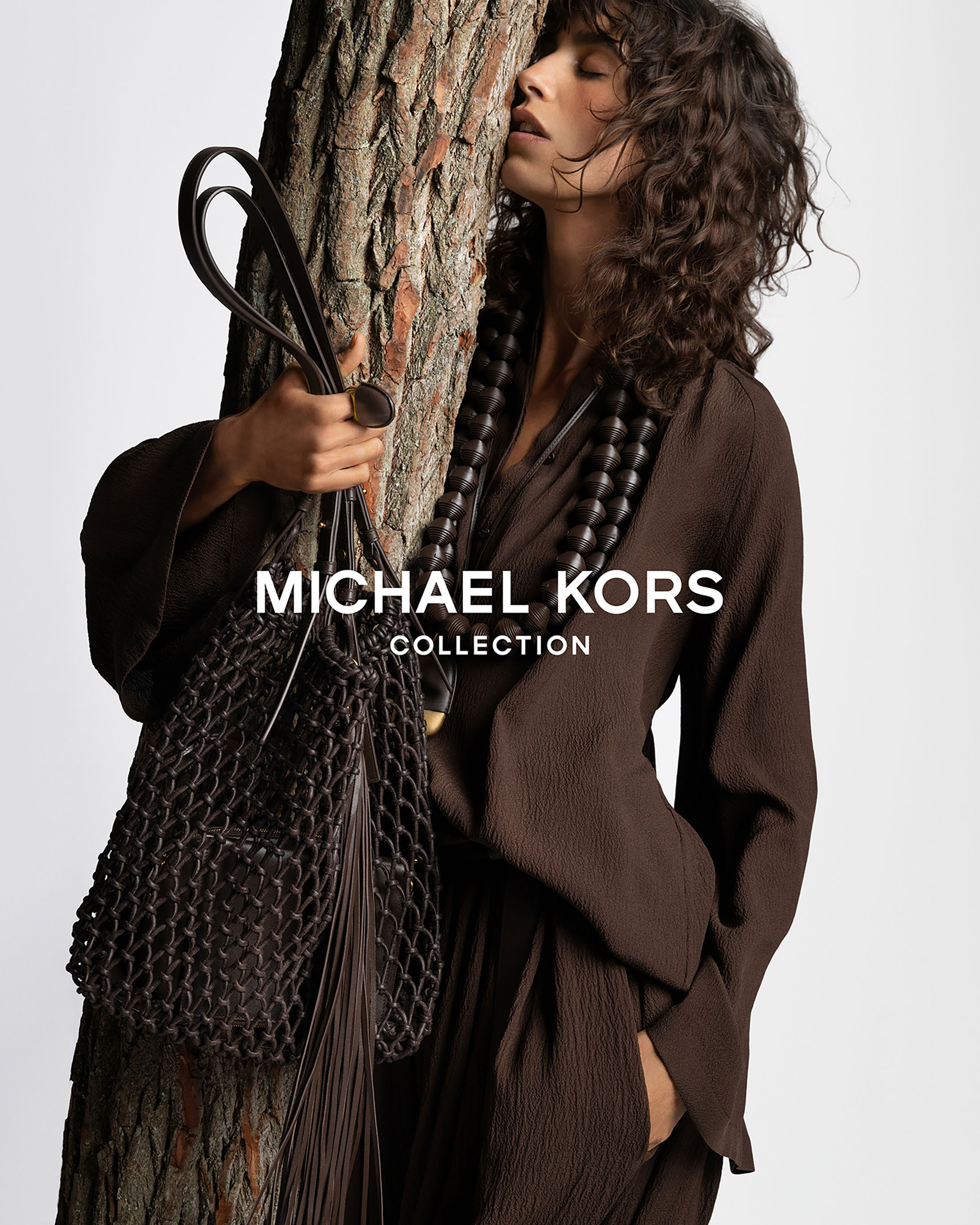 Brunette woman in brown outfit leans against tree, holding Michael Kors net bag. Dark beaded necklace is prominent.