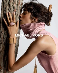 Model Mica Argañaraz wearing a pink turtleneck and statement ring leaning against a tree trunk for Michael Kors.