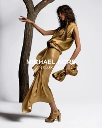 Model Mica Arganaraz wearing a metallic gold dress and heels holding a clutch next to a tree trunk for Michael Kors.