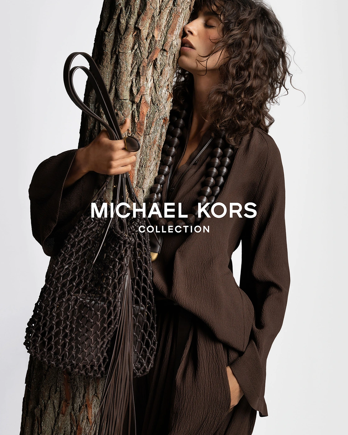 Model Mica Argañaraz leans against a tree trunk holding a fringed bag for the Michael Kors Spring 2026 ad campaign.