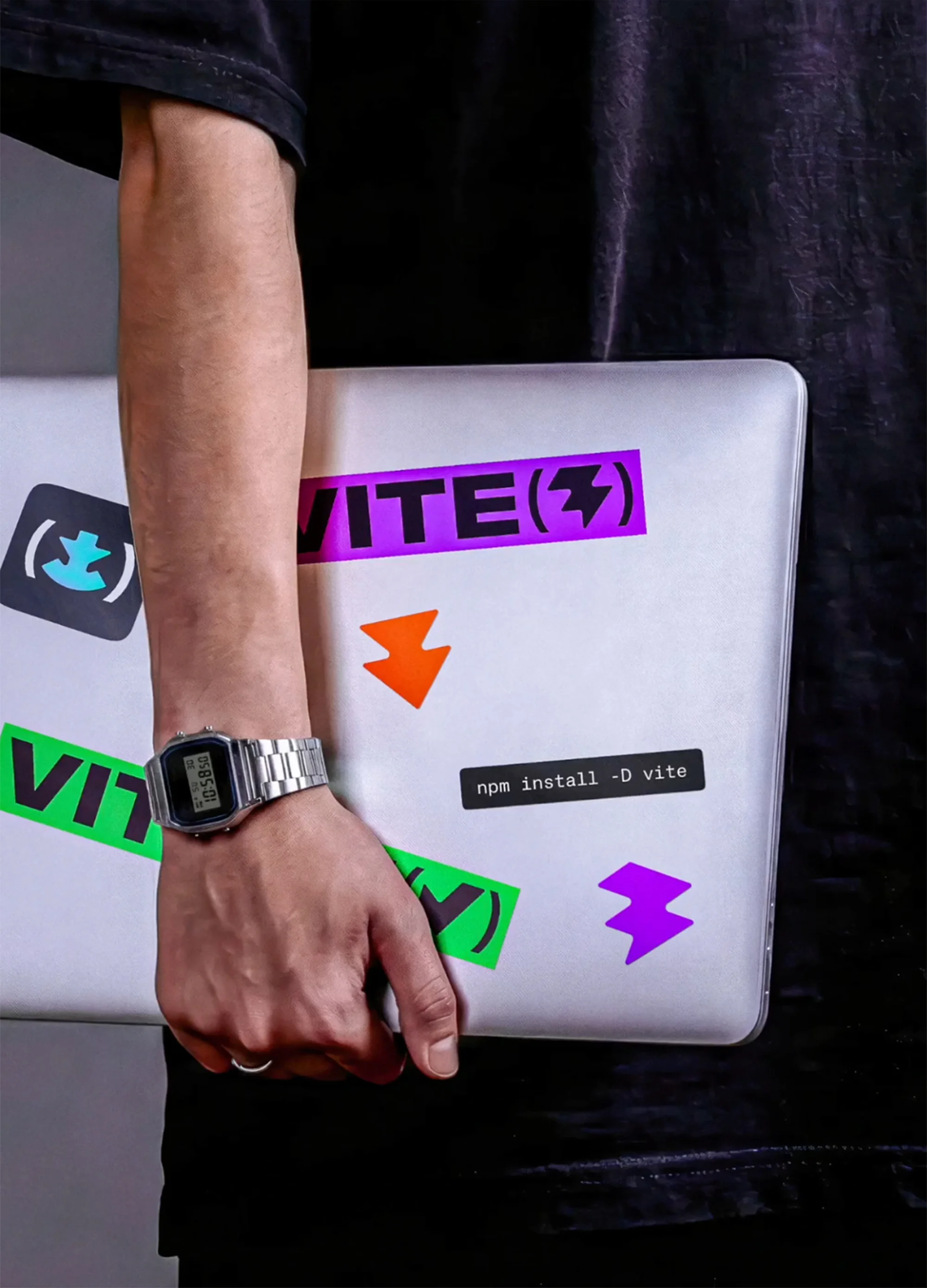 Person carrying a laptop with stickers, wearing a digital watch, and dressed in a black t-shirt.