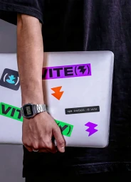 Close-up of a person holding a silver laptop covered in colorful stickers, wearing a black t-shirt and a digital metal watch.