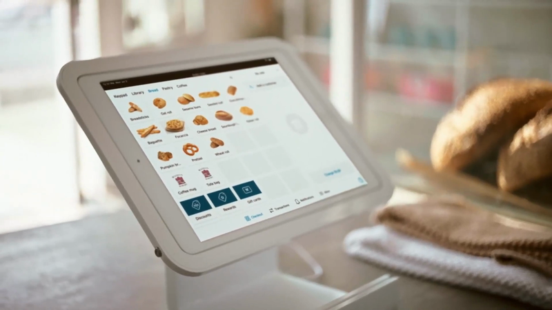Digital ordering system displayed on a tablet with bread options at a cafe, with baked goods blurred in the background.