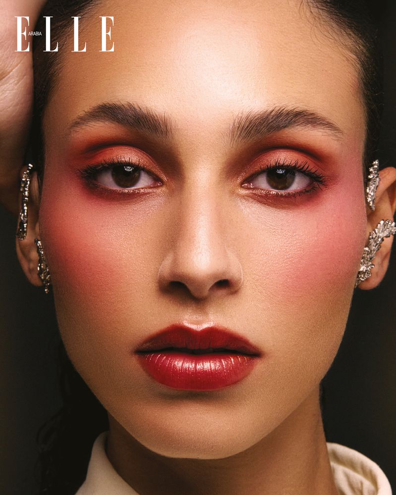 Close-up portrait of a woman with dramatic red makeup and decorative ear cuffs, against a dark background; Elle magazine logo is visible.