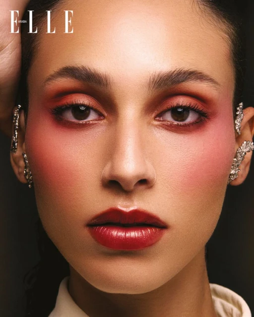 Close-up portrait of a woman with dramatic red makeup and decorative ear cuffs, against a dark background; Elle magazine logo is visible.