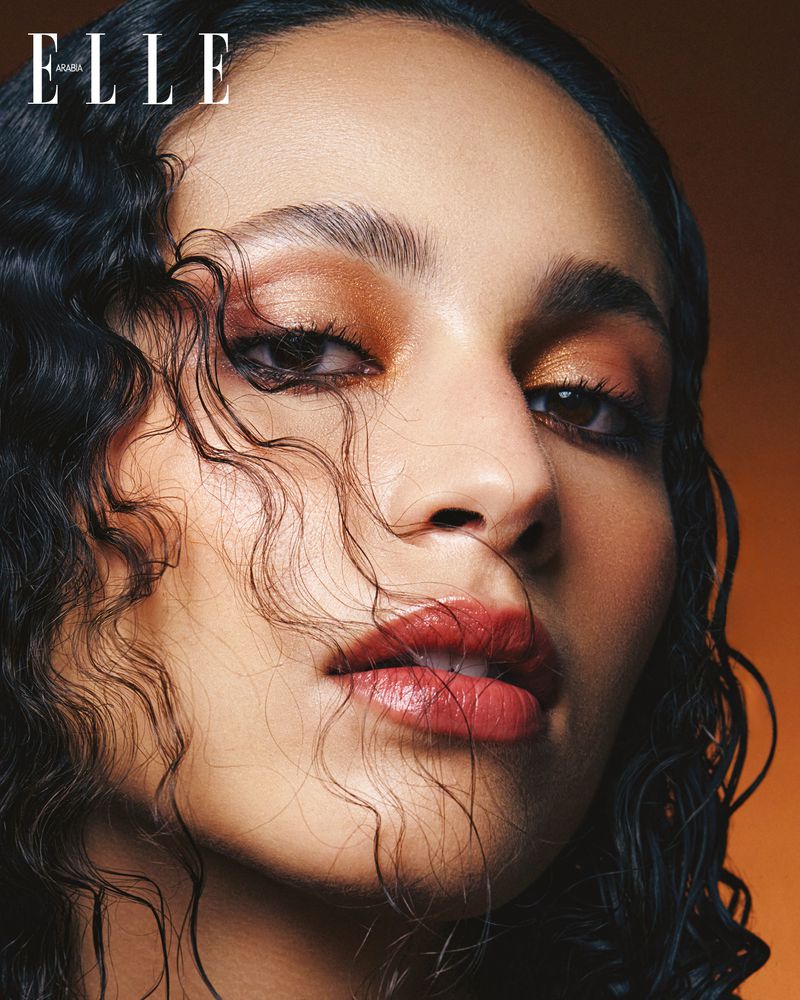 Close-up portrait of an adult woman with dark, curly hair and bronze eye shadow looking at the camera against an orange gradient background.