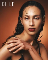 Close-up portrait of a woman with wet dark hair and deep red lipstick touching her shoulder against a warm amber gradient background.