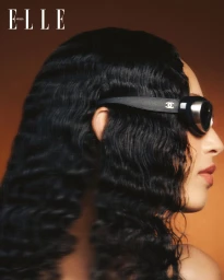 Model in profile wearing dark sunglasses with long wavy black hair against a warm gradient background for Elle Arabia Chasing light