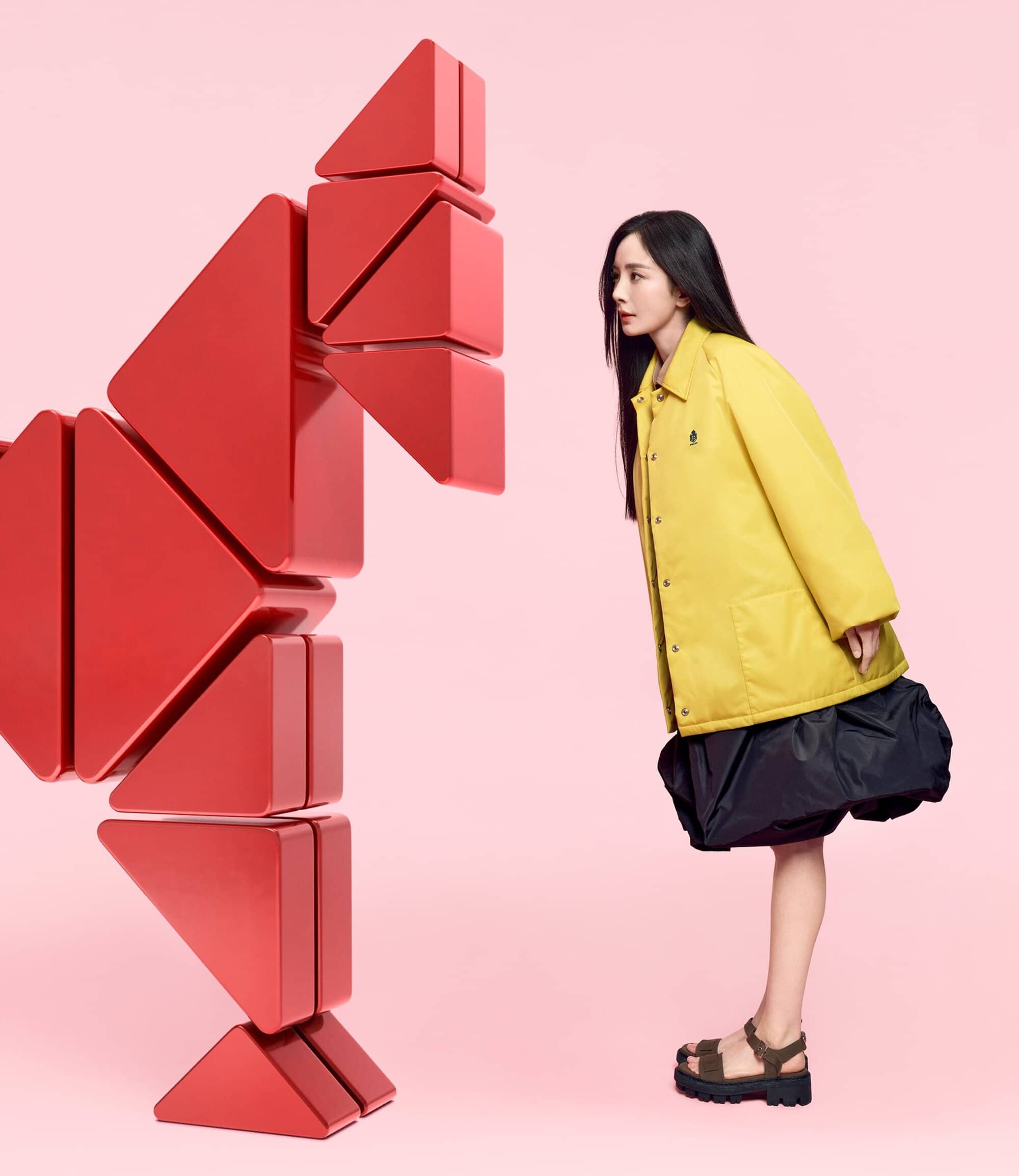 Adult woman in bright yellow jacket gazes at a geometric red object in a pink studio. Fashion editorial style, eye-level angle.