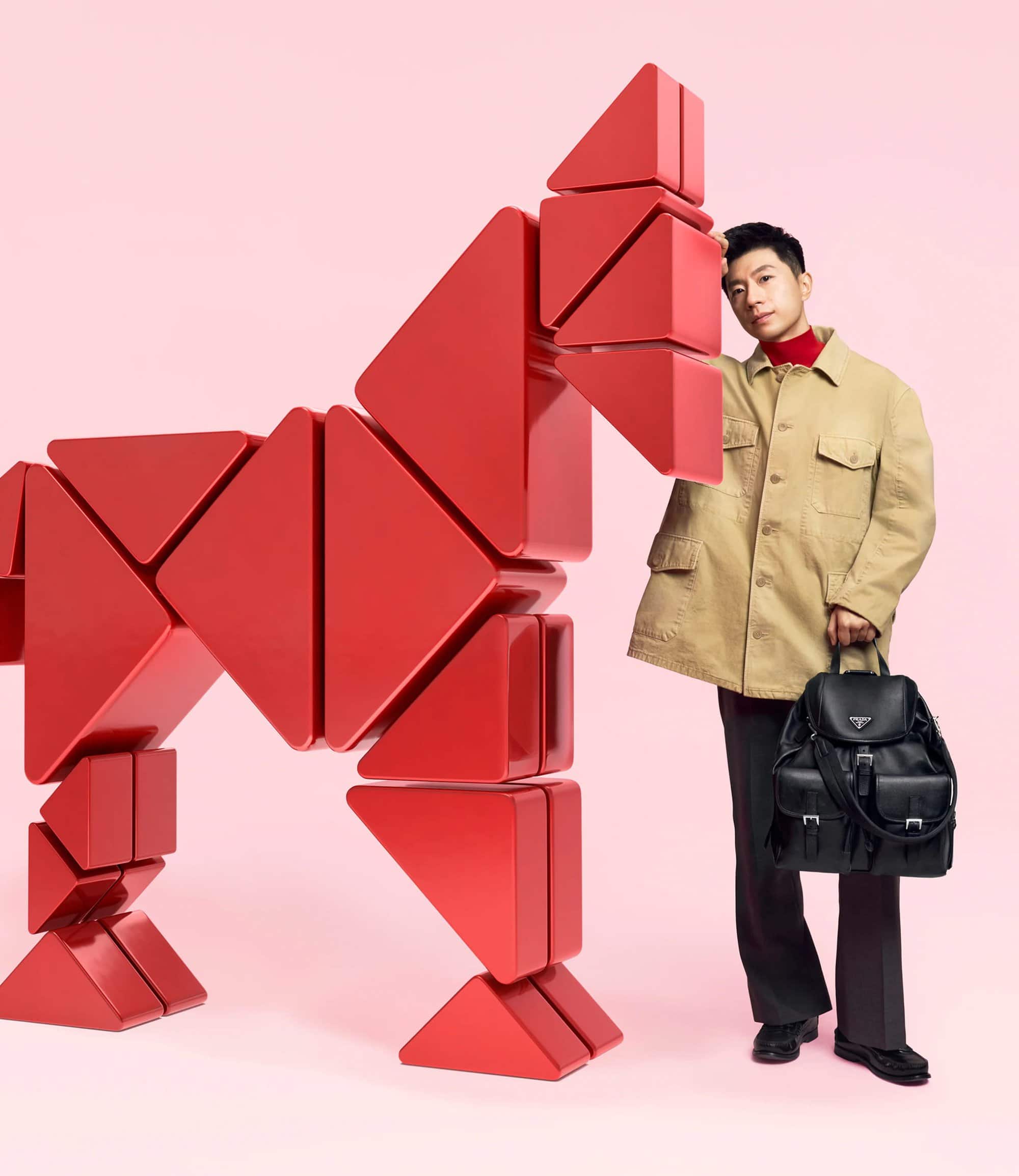 An Asian man in a tan jacket poses next to a red geometric sculpture of a horse, holding a black backpack in a studio setting.