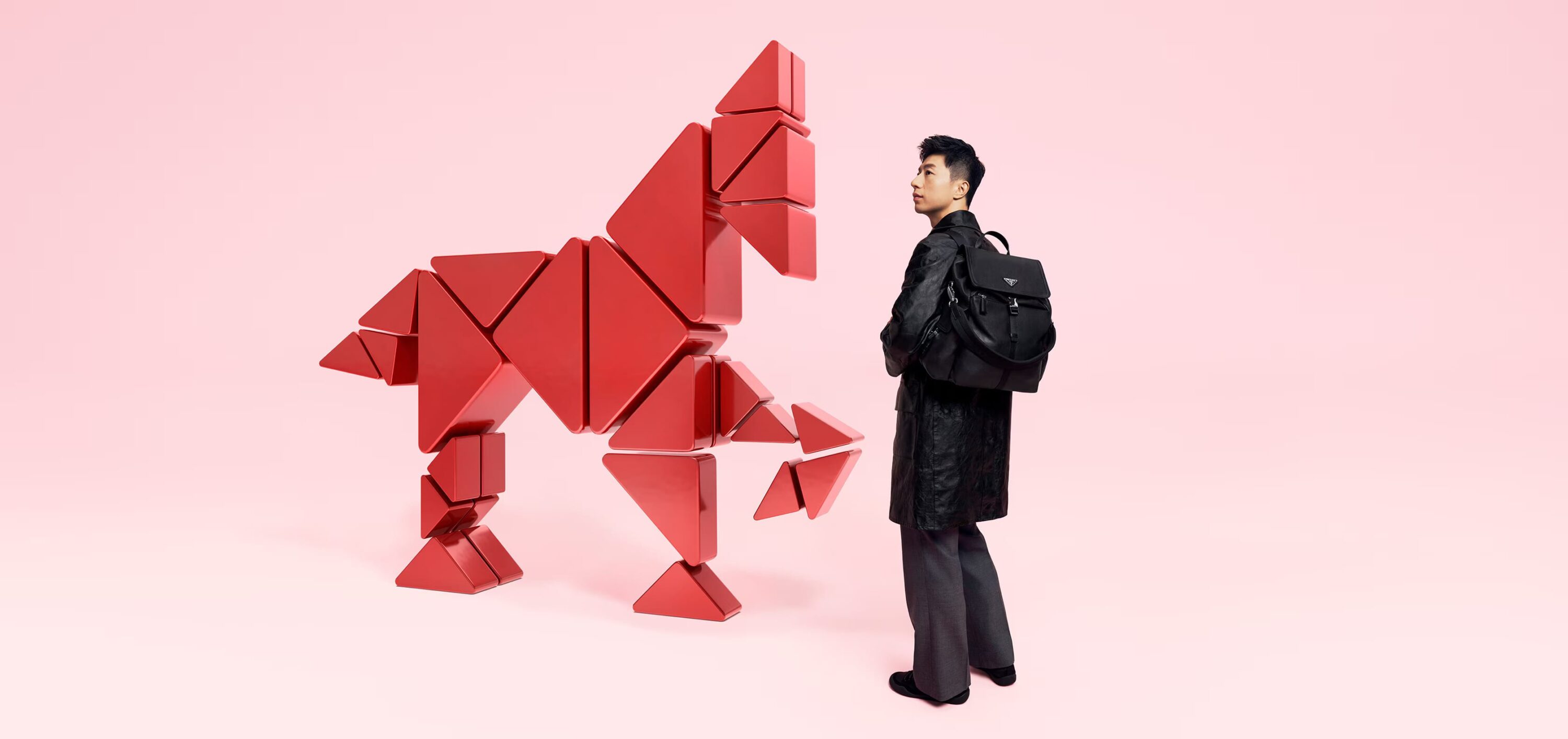 An adult man in a dark coat with a backpack stands near a sculpture of a horse constructed from red triangular blocks against a pink backdrop.