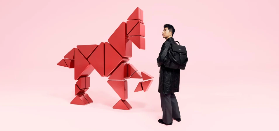 An adult man in a dark coat with a backpack stands near a sculpture of a horse constructed from red triangular blocks against a pink backdrop.