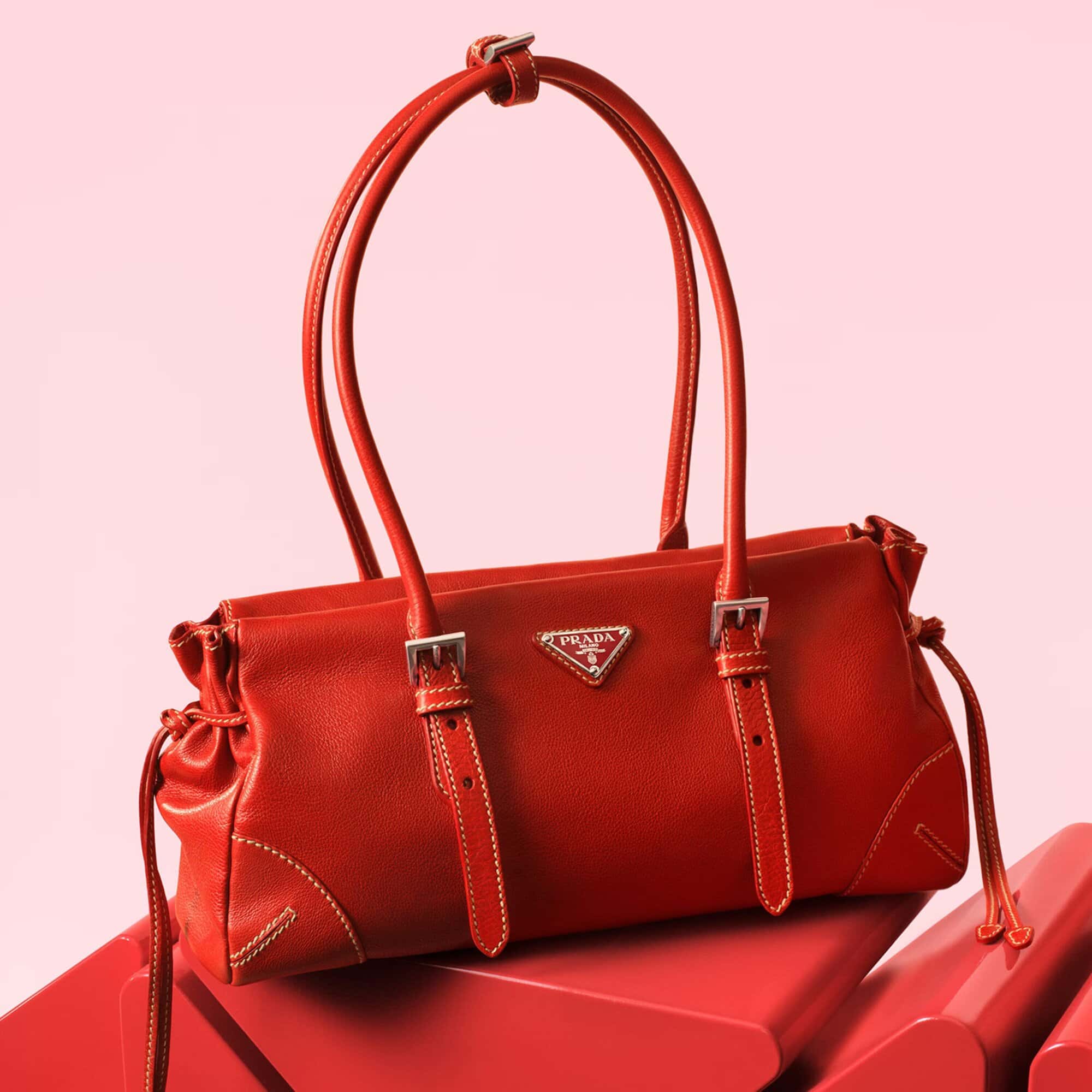 Studio shot of a red Prada handbag with double handles and silver hardware, staged on stacked geometric forms against a pink backdrop.