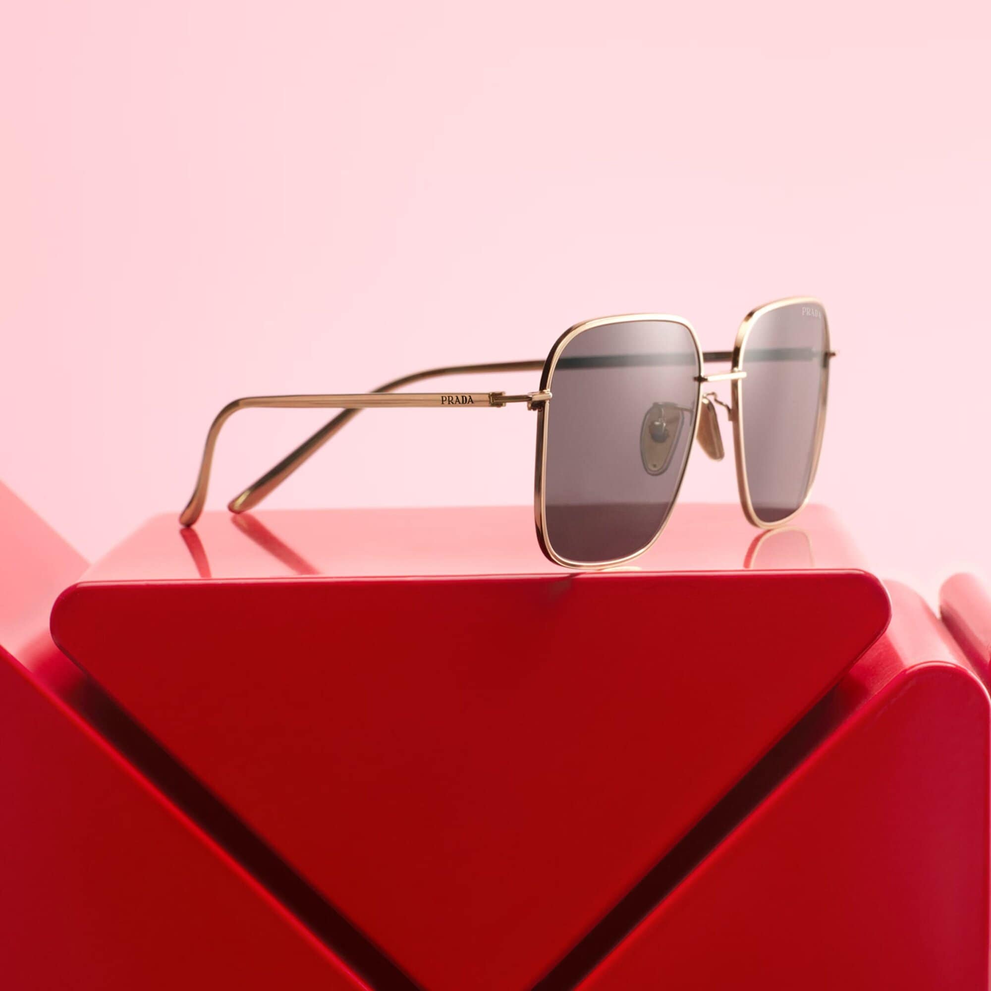 Square, gold-framed Prada sunglasses with grey lenses displayed on a geometric red pedestal against a pastel pink background.
