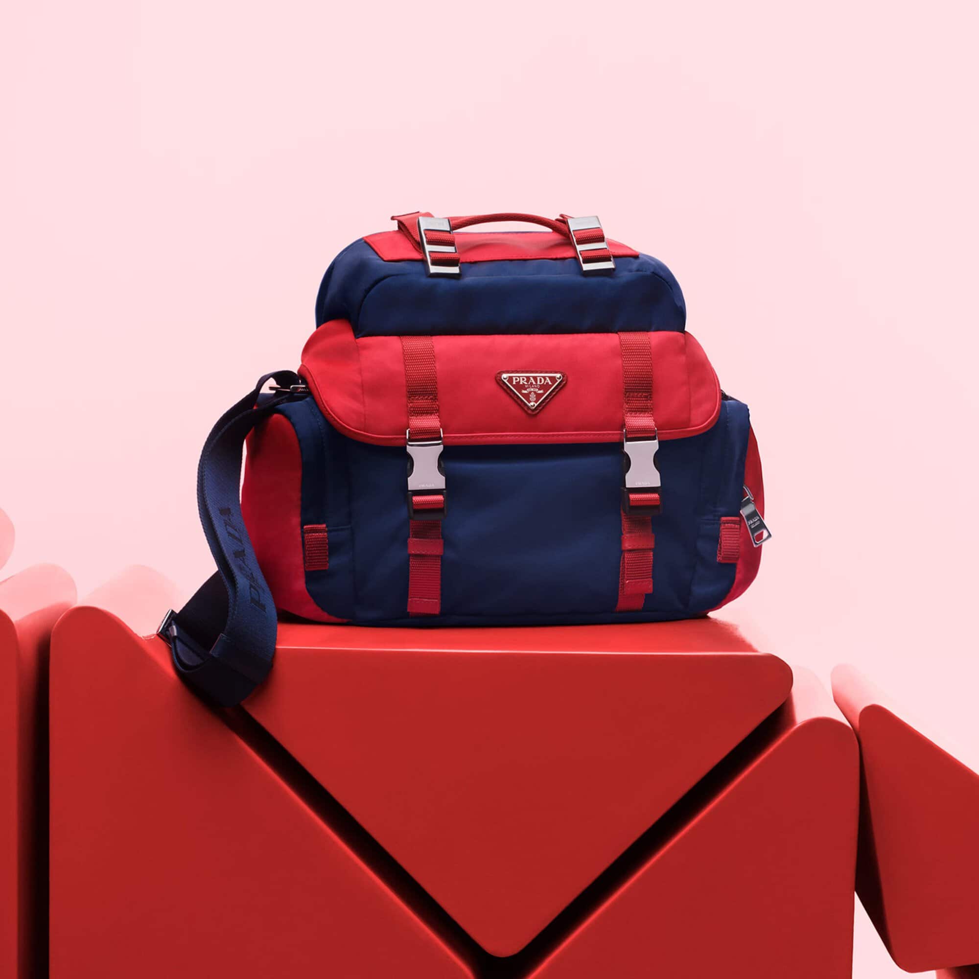 Prada backpack in blue and red on a red geometric platform against a pink background. Fashion product shot with studio lighting.