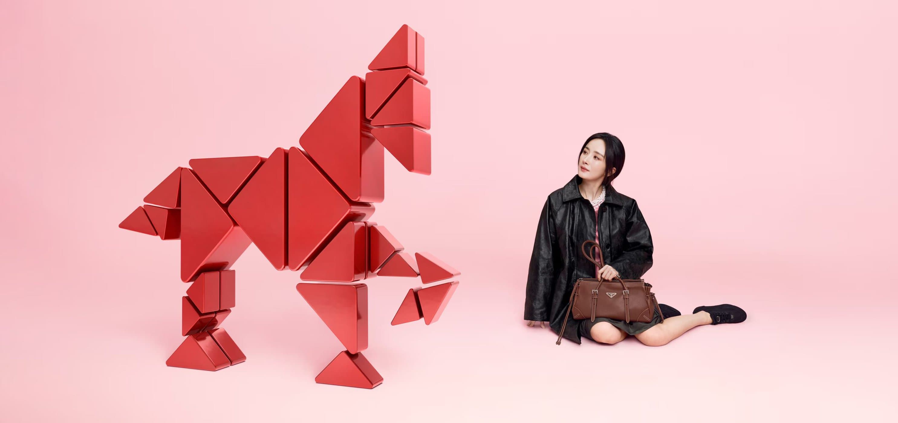 Young woman in a black jacket sitting with a brown purse next to a red geometric horse sculpture against a pale pink backdrop.