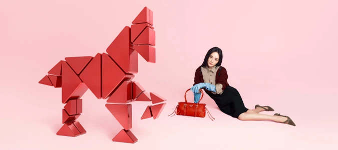 Woman in dark hair and layered cardigan poses beside a large geometric red horse sculpture against a soft pink backdrop.