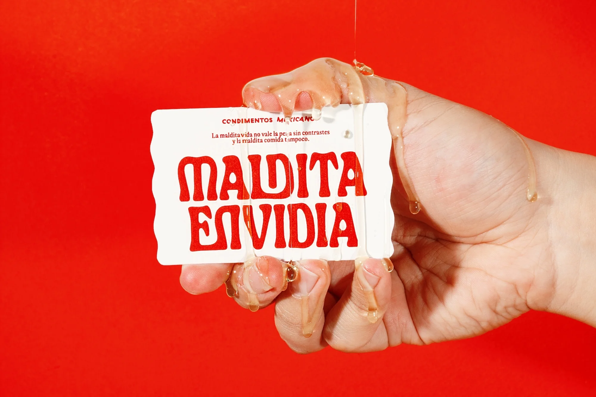 A hand holds a card with the text 'Maldita Envidia' dripping with a clear substance, set against a vibrant red background.