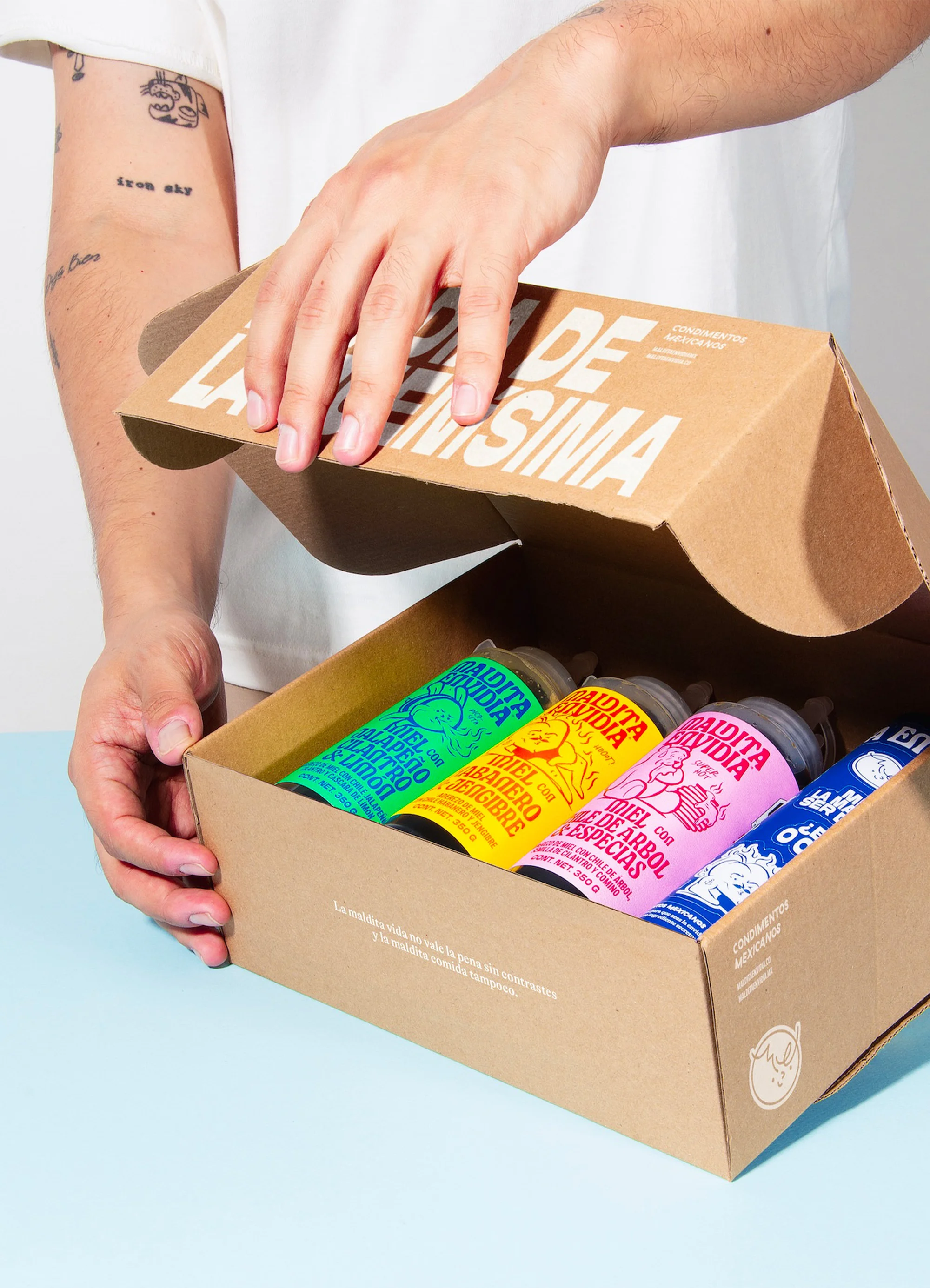 Person opening a cardboard box containing four colorful bottles, with white background and soft lighting, showcasing product packaging.