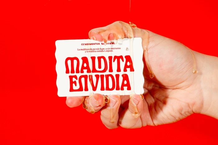 Hand holding a white card with bold red Maldita Envidia typography, covered in dripping honey against a solid red background