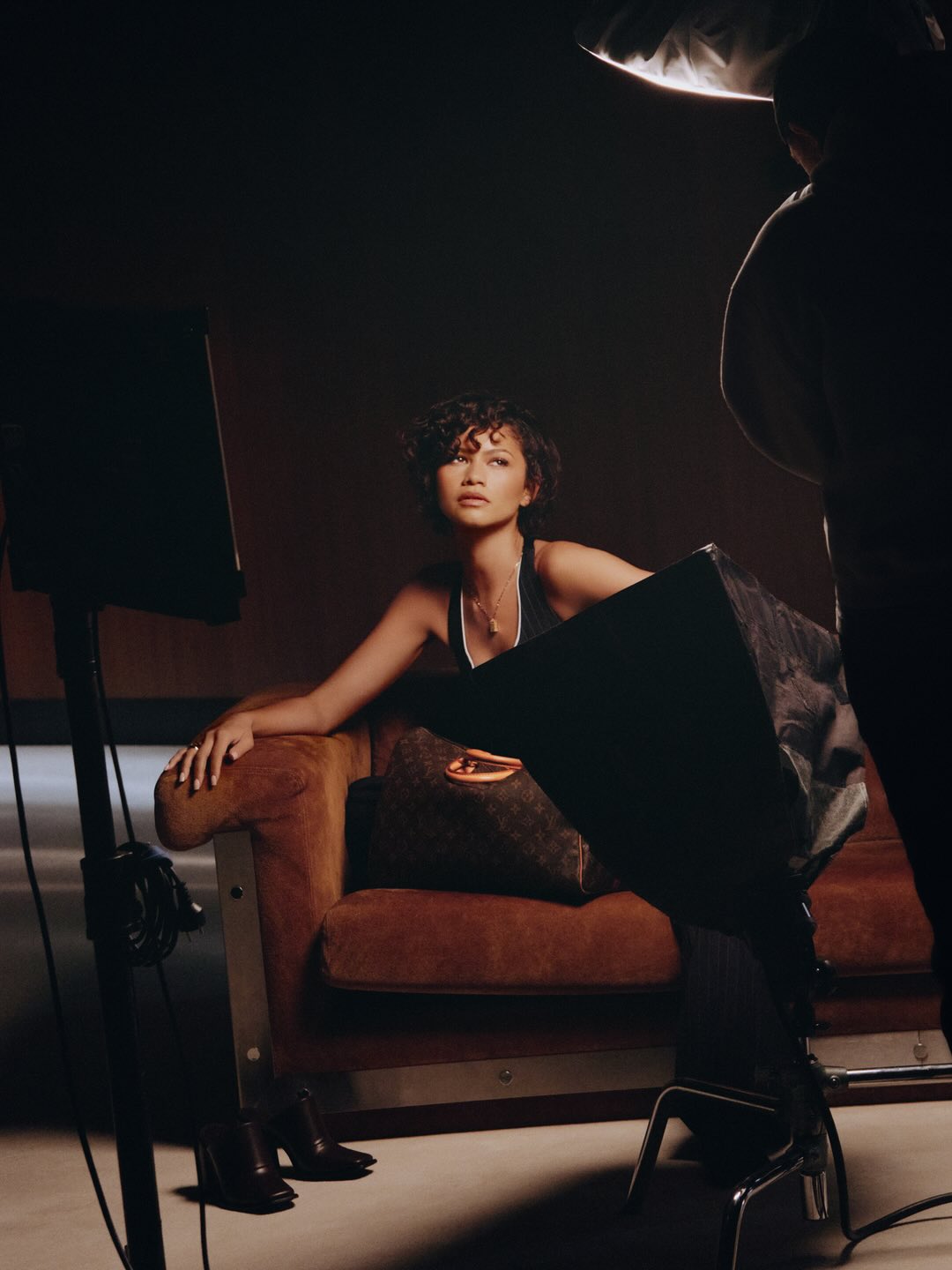 A woman sits on a brown sofa in a photo studio setting, accompanied by a Louis Vuitton bag, with studio lighting equipment surrounding her.