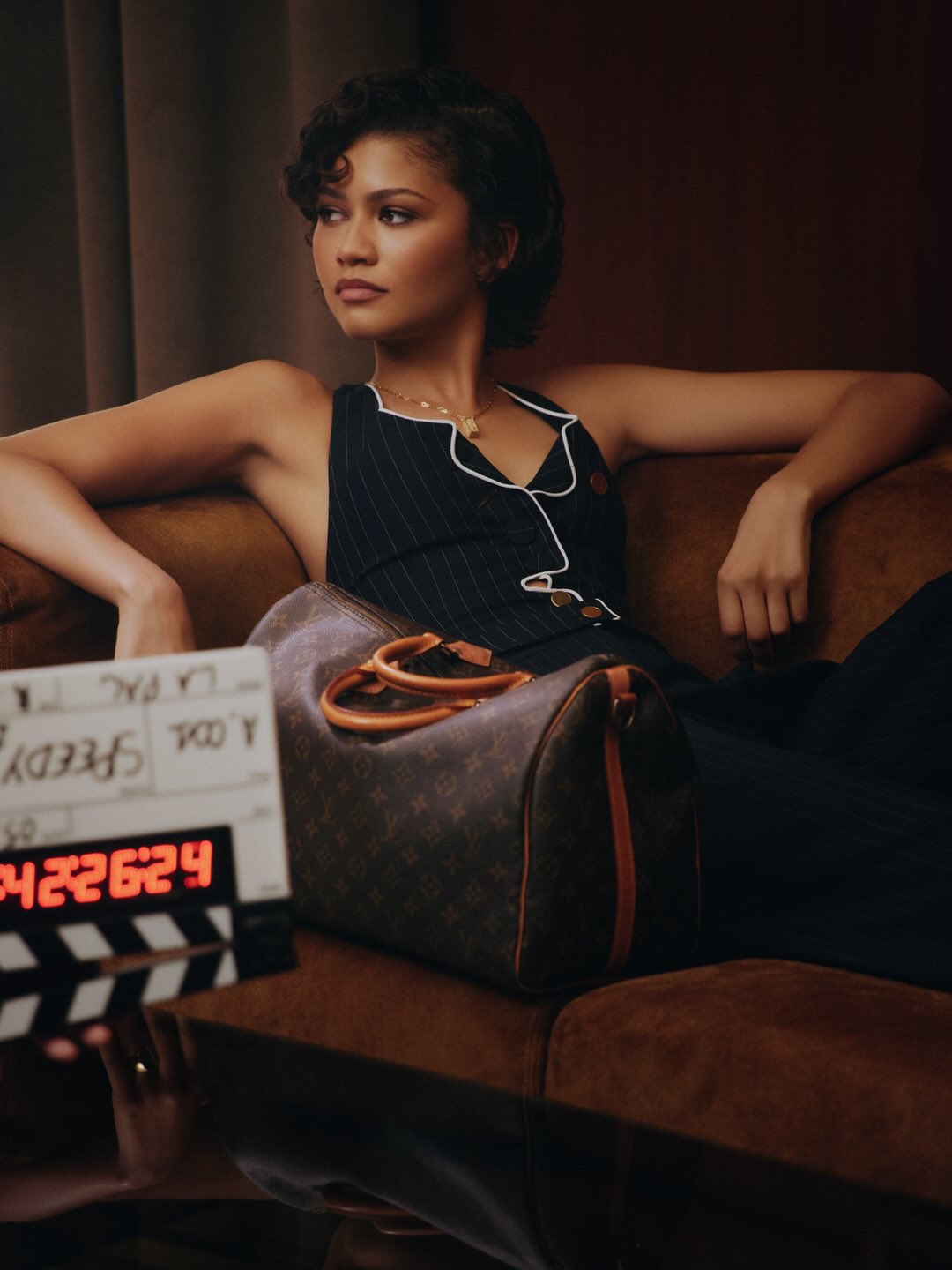 Zendaya poses on a brown sofa with a Louis Vuitton bag and clapperboard, wearing a dark pinstripe sleeveless dress with white trim.