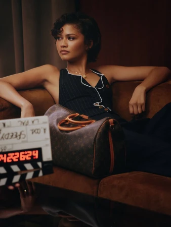 Zendaya poses on a brown sofa with a Louis Vuitton bag and clapperboard, wearing a dark pinstripe sleeveless dress with white trim.