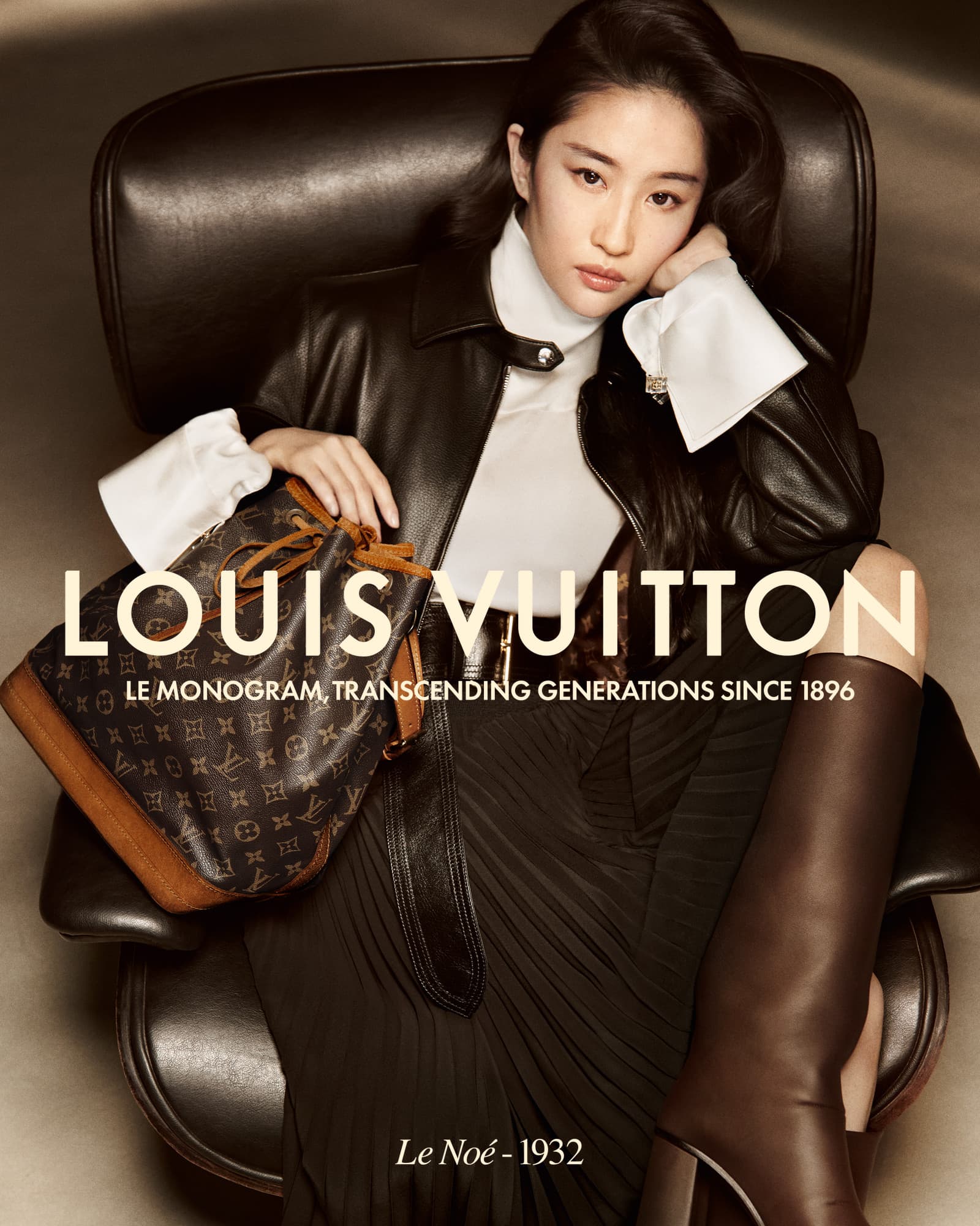 Asian woman in a dark leather jacket poses with a Louis Vuitton purse while sitting in a black leather chair in a studio.