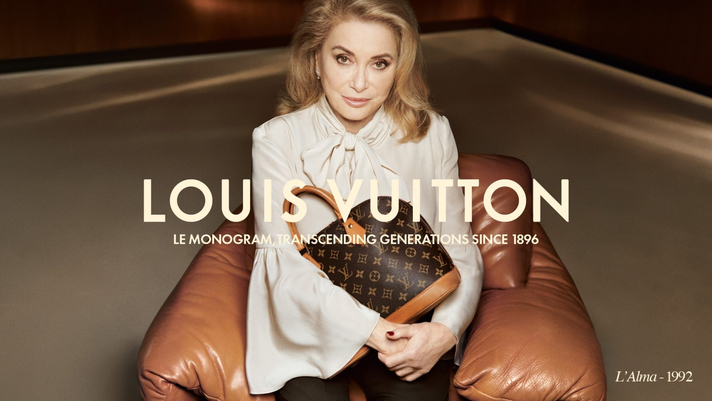 Catherine Deneuve sits with a Louis Vuitton monogram bag, stylish clothing, and minimal background in this luxury brand fashion campaign.