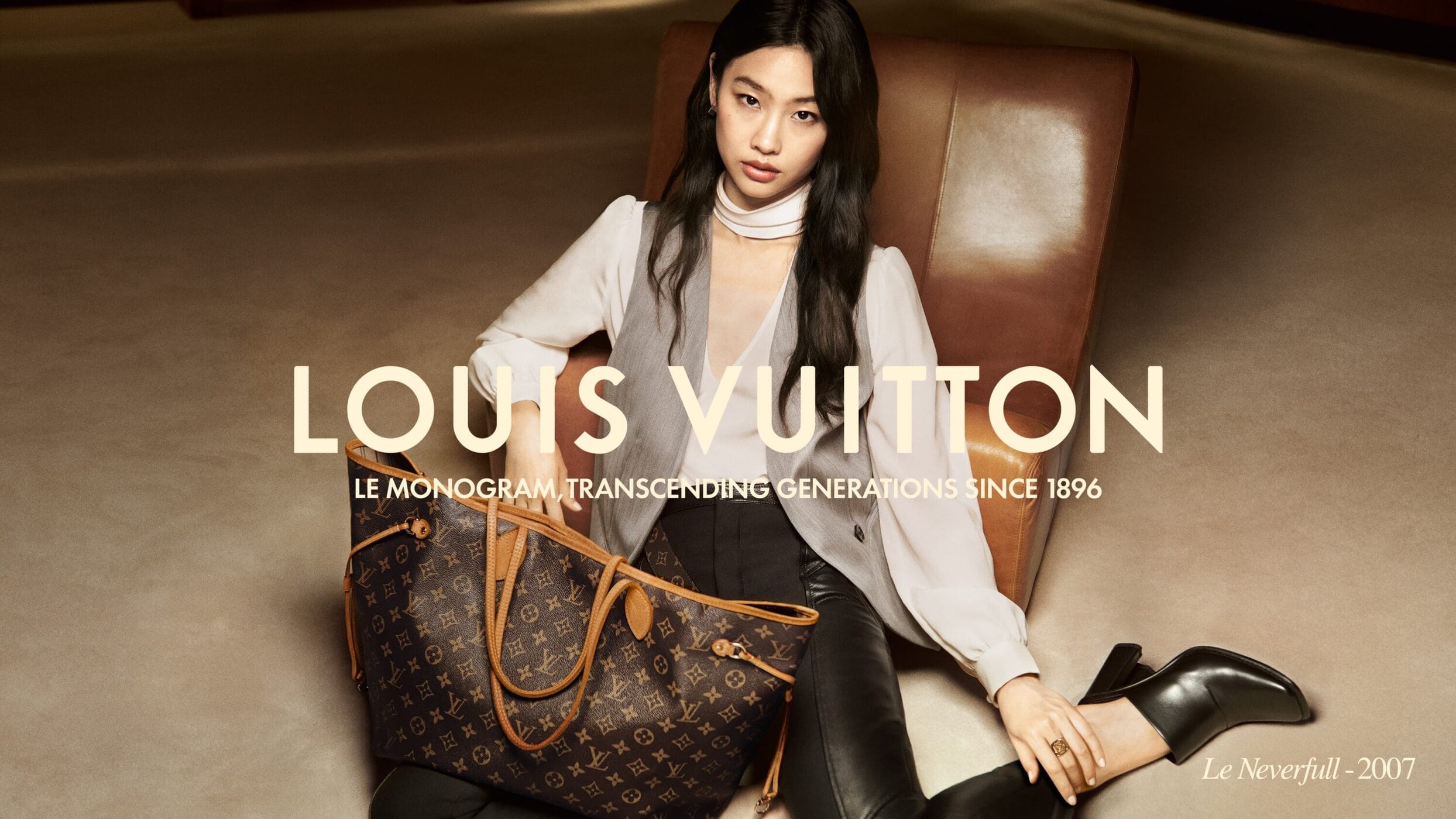 An Asian female model in a gray vest and black pants sits in a warm-toned armchair with a Louis Vuitton handbag on the floor.